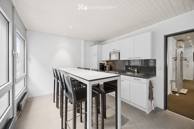 Modern kitchen with white cabinets, a black countertop, a bar table with bar stools, and a sink with a faucet. The space is bright with large windows and recessed lighting.