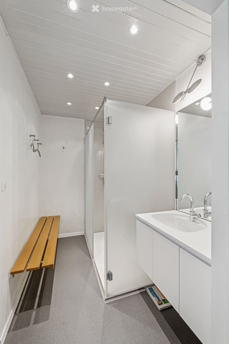 Modern bathroom with white walls, a double sink, a shower, and a wooden bench. The ceiling features recessed lighting, and the floor is carpeted.