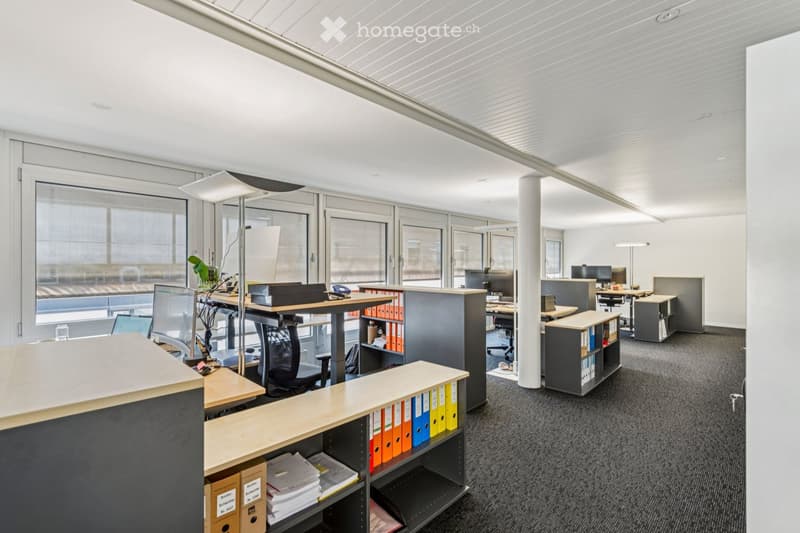 Modern office space with desks, chairs, computers, and filing cabinets arranged around a central area, featuring large windows and a sleek design.