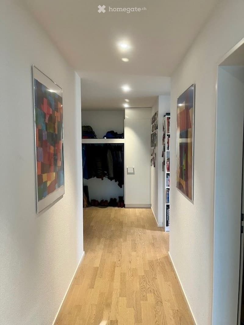 A hallway with a wooden floor, a closet with hanging clothes, a wall with two paintings, and an open door.