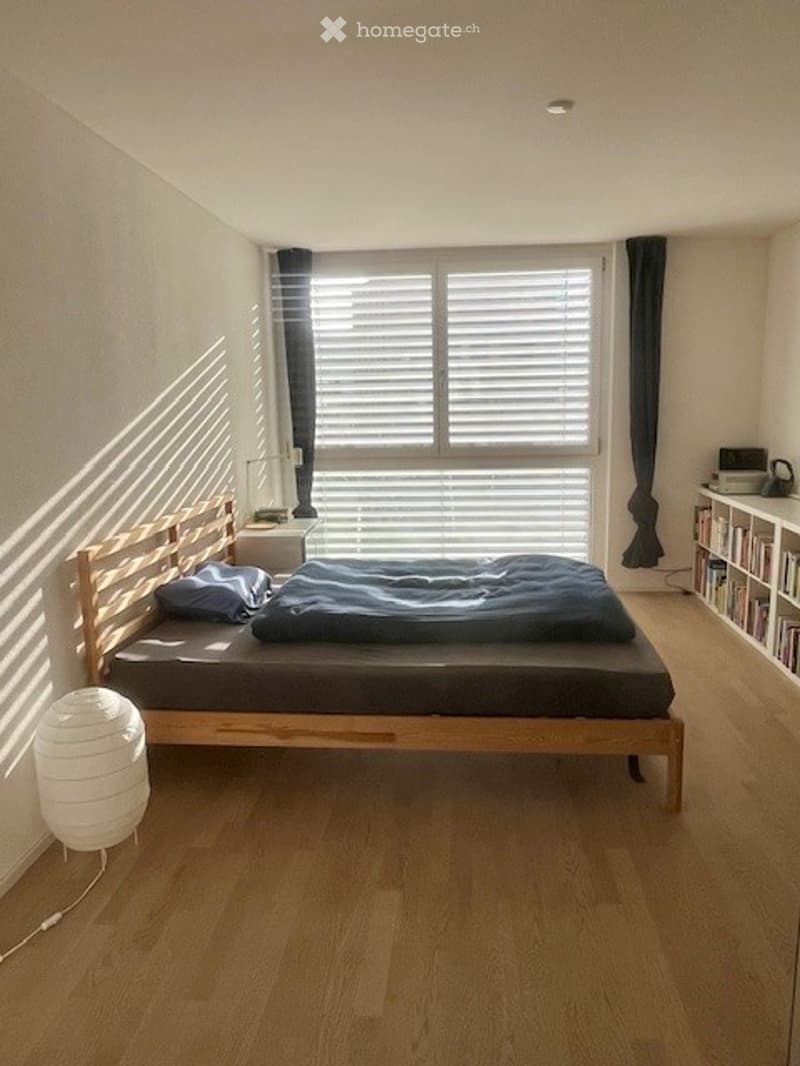 wooden bed, 2 pillows, lamp, nightstand, large window with blinds, bookcase, books, floor lamp
