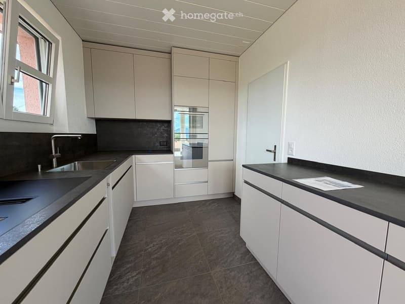 Modern kitchen with white cabinets, black countertops, stainless steel sink, and built-in oven. Dark tiled floor and large window provide natural light.
