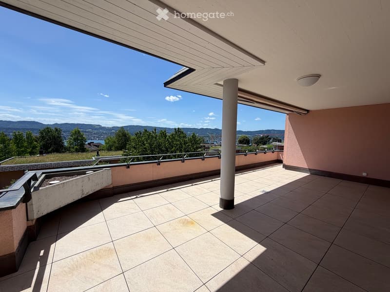 Balcony with tiled floor, metal railing, and a small planter, overlooking a scenic landscape with trees and mountains.