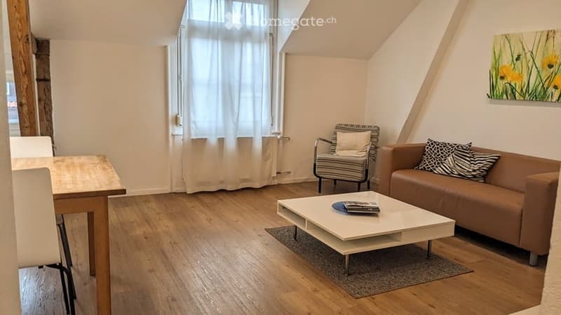 Living room with wooden flooring, large window with white curtains, brown sofa, white coffee table, striped armchair, and small table with chairs.