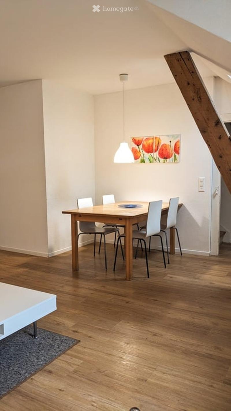 Bright dining area with wooden floor, white walls, and a wooden dining table surrounded by white chairs. A pendant light hangs above, and a colorful painting of tulips adorns the wall.