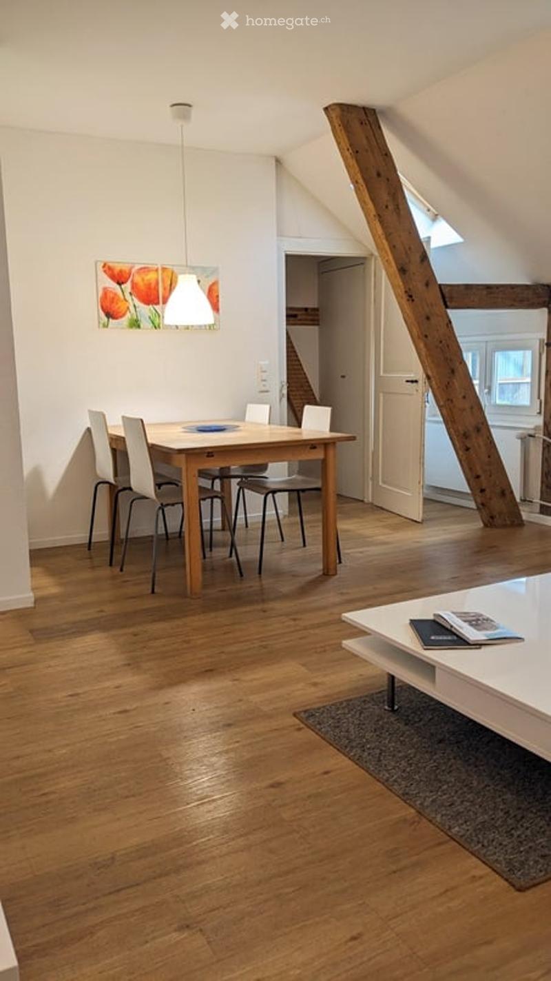 Bright, open-plan living and dining area with wooden flooring, white walls, and exposed wooden beams. Features a dining table with chairs, a coffee table with books, and a picture frame on the wall.