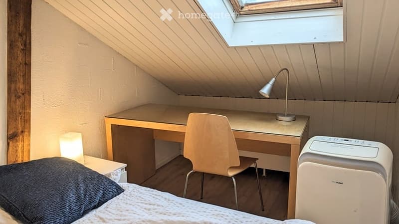 Cozy attic bedroom with a slanted ceiling, featuring a bed, desk, chair, and a small lamp on the desk.