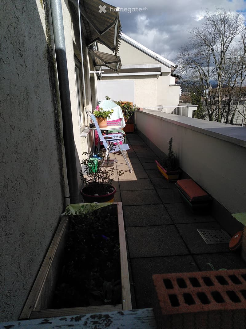 Balcony with tiled floor, potted plants, small table and chairs, and a view of the building and surrounding area.