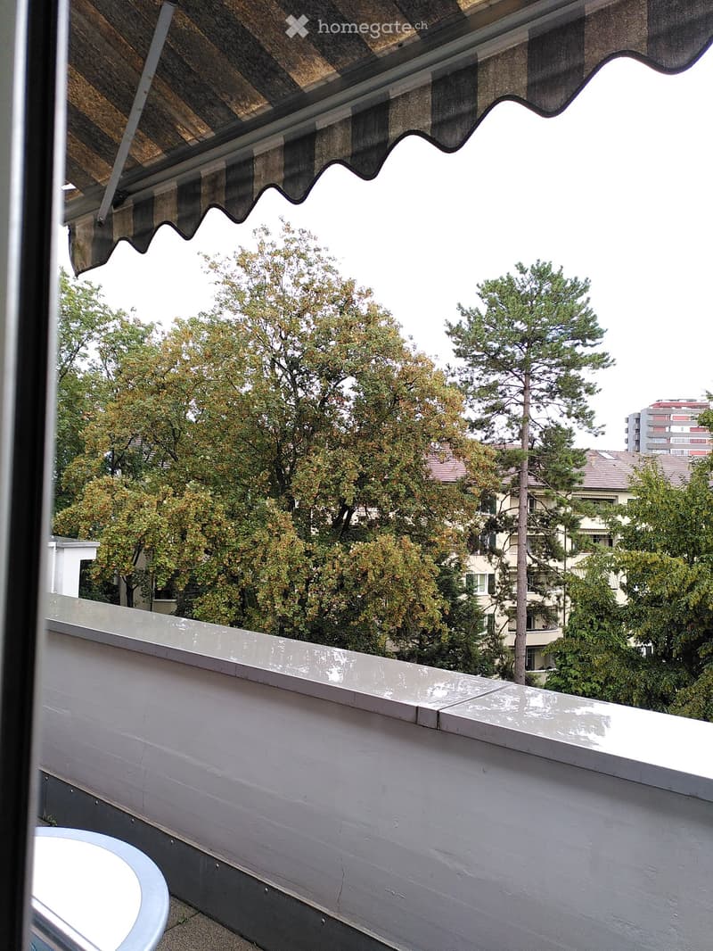 Balcony with a white railing, a round table, and a view of trees and buildings.