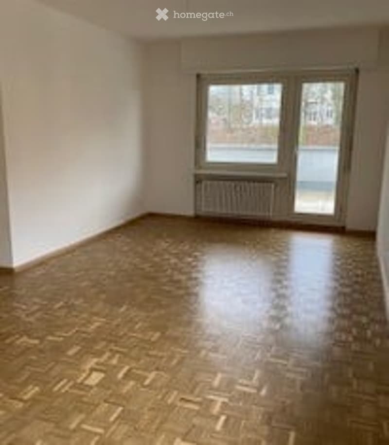 Completely empty room with wooden herringbone flooring, white walls, large window with radiator below, and glass door leading outside.