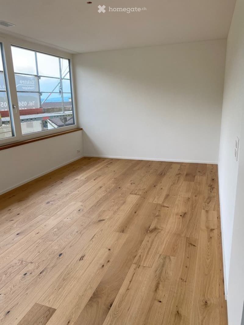 Empty room with light wooden flooring and white walls, featuring large windows with a view of the outside.