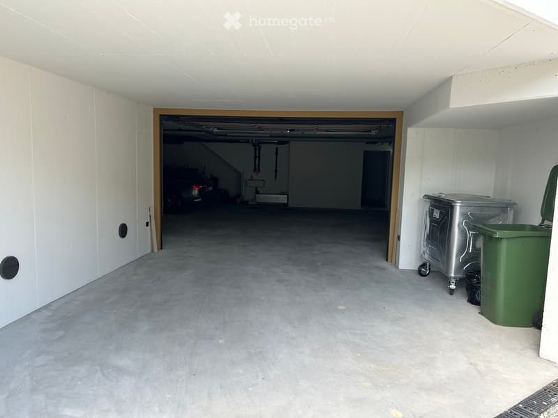 Empty garage with concrete floor, white walls, and a large open doorway leading to another space. A green trash bin and a metal cart are visible on the right side.