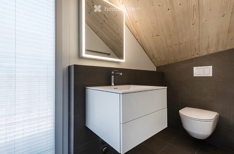 Modern bathroom with a white sink, sleek mirror, and toilet. Dark grey tiles on walls and floor, wooden slanted ceiling.