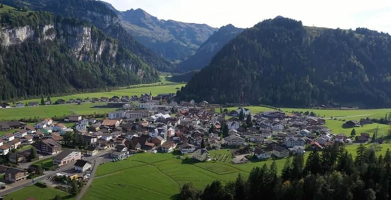 Aerial view of a small village nestled in a valley surrounded by mountains, with houses, green fields, and a church visible.