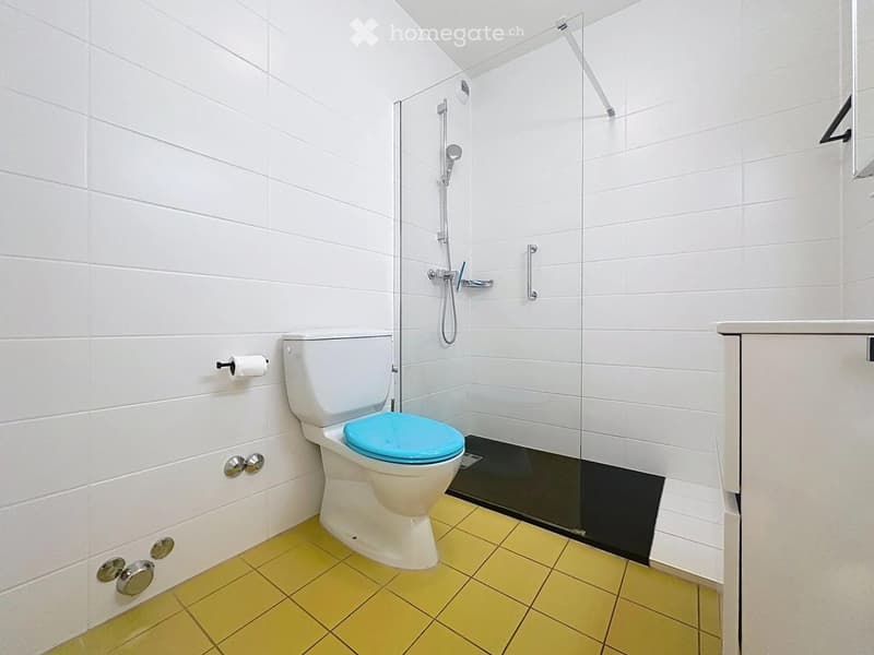 Modern bathroom with white tiled walls, yellow tiled floor, toilet with blue seat, glass-enclosed shower, and toilet paper holder.