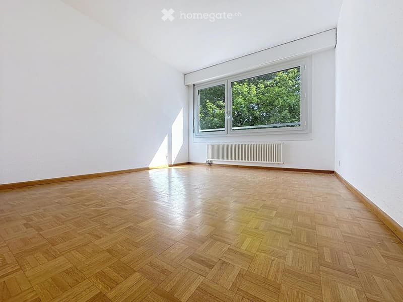 Empty room with wooden parquet flooring, white walls, and a large window with white frame. Natural light streams in, illuminating the space.