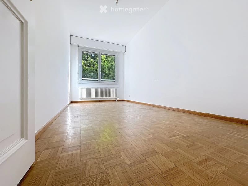Empty room with white walls, wooden herringbone flooring, and a single window with white frame and radiator below.