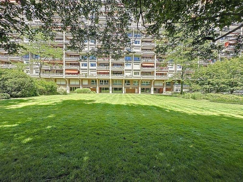 A large, well-maintained lawn in front of a multi-story residential building with numerous balconies and windows, surrounded by trees and greenery.