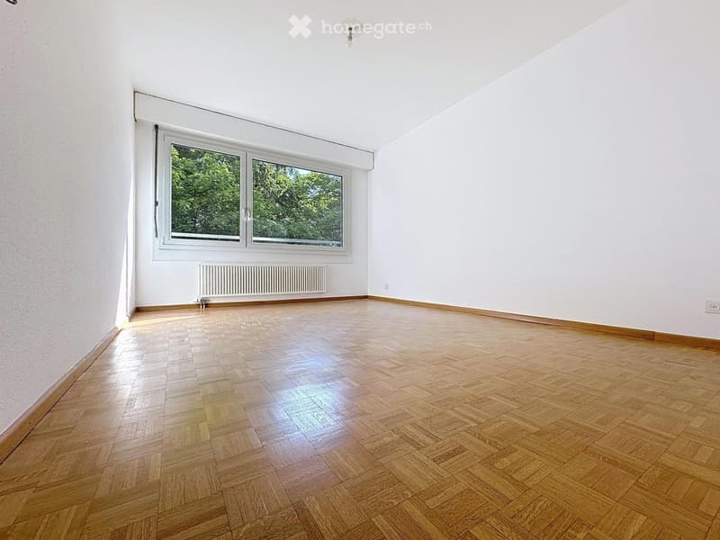 Empty room with wooden parquet flooring, white walls, large window with white frame, radiator, and visible electrical outlets.