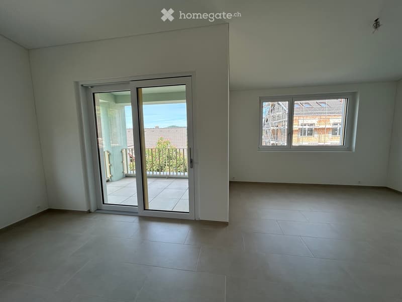 Empty room with tiled flooring, white walls, large sliding glass door leading to a balcony, and a window overlooking a construction site.