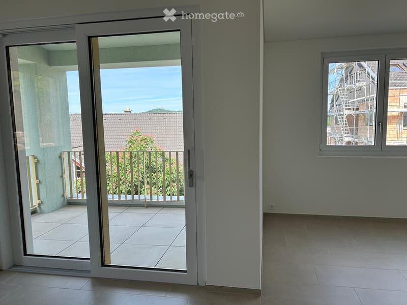 Empty room with tiled flooring, large sliding glass doors leading to a balcony with a view of the roof and greenery.