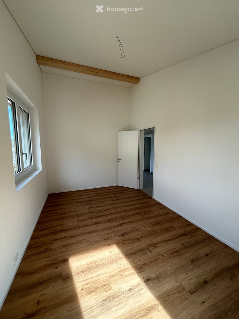 Empty room with wooden flooring, white walls, and a window letting in natural light. The room features a door leading to another space.