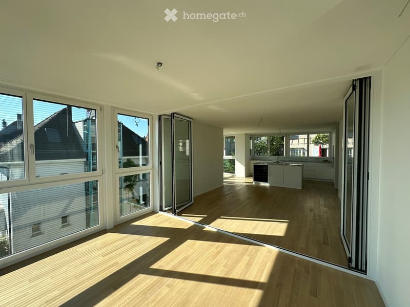Bright, empty room with wooden flooring, large windows, and sliding glass doors leading to another space.