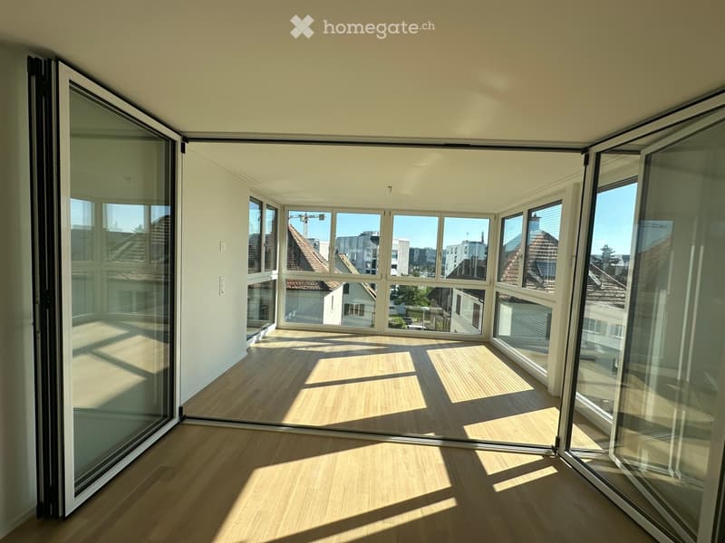 Bright, empty room with large glass windows and sliding doors, wooden flooring, and natural light.