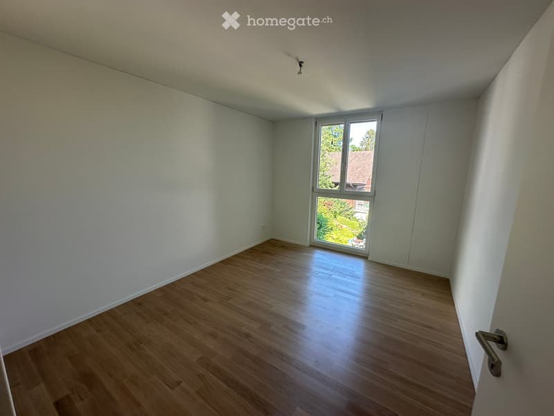 Empty room with light-colored wooden flooring and white walls, featuring a large window with a view of greenery and a neighboring building.