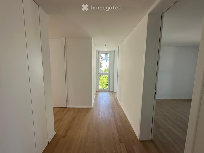 Empty hallway with wooden flooring, white walls, and a window at the end letting in natural light.