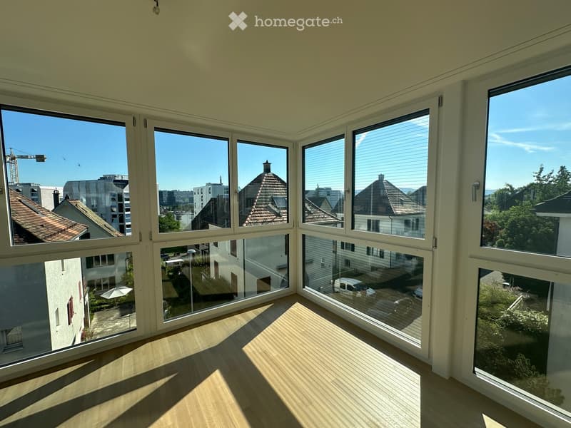 Empty room with large windows offering a view of the cityscape and surrounding buildings. Light wooden flooring and white walls.