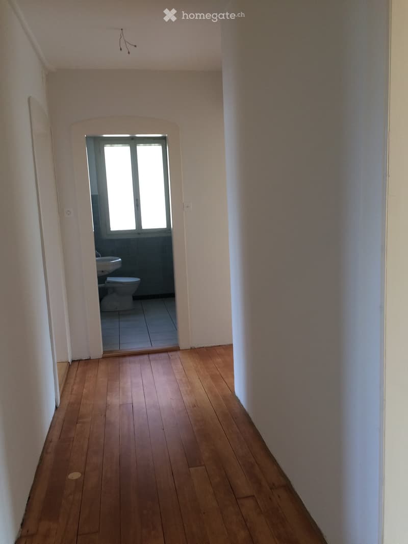 Empty hallway with wooden flooring and white walls, leading to a small bathroom with a toilet and sink.