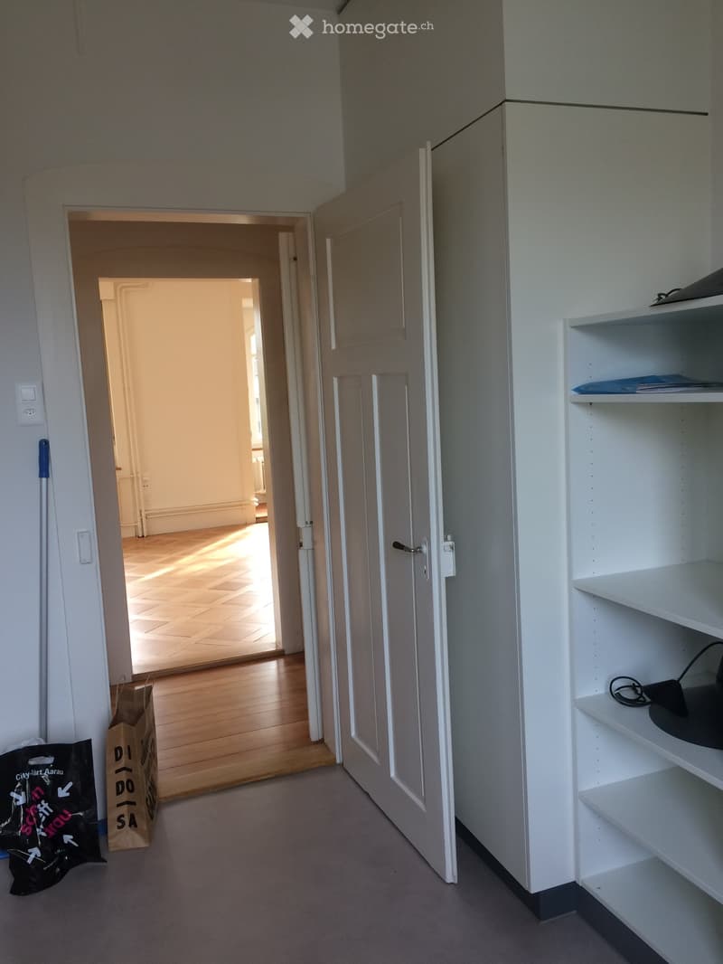 Empty hallway with light-colored walls, carpeted floor, and open doors leading to another room with wooden flooring.