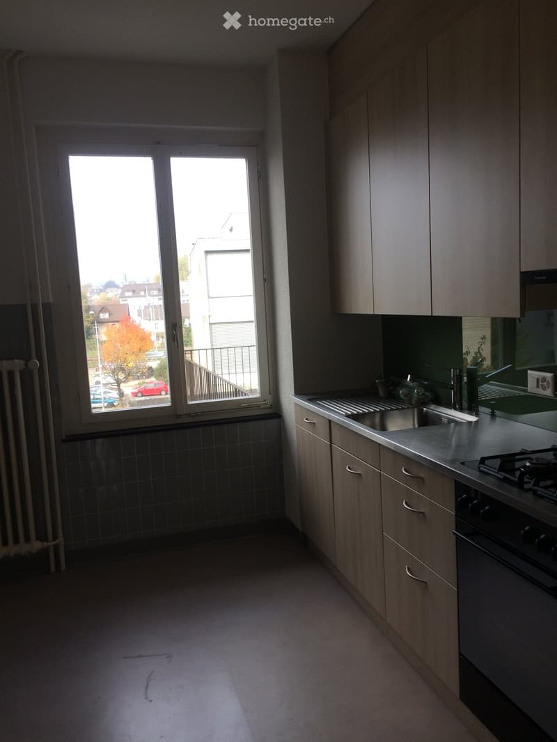Modern kitchen with light wood cabinets, stainless steel sink, gas stove, and oven. Large window offers a view of the street and autumn foliage.