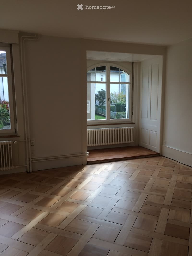 Empty room with wooden herringbone flooring, large arched window, radiator, and white walls.
