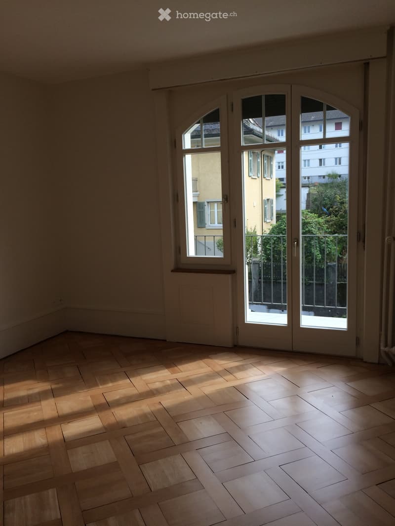 Empty room with wooden flooring and large windows, allowing natural light to fill the space. The room features a balcony accessible through the windows.
