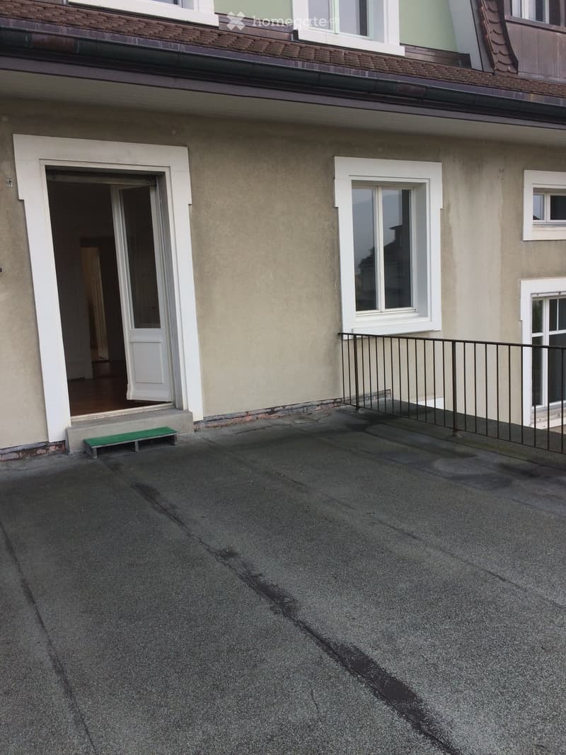 Roof terrace with concrete surface, metal railing, green bench, and open doorway to the building.