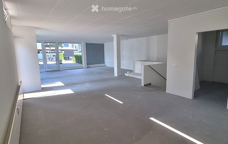 Empty room with white walls, concrete floor, large glass doors leading to an outdoor area, and a small white table near the entrance.