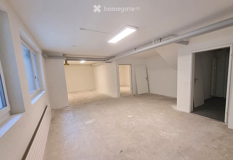 Empty room with white walls, concrete floor, large window, and several doors leading to other spaces.