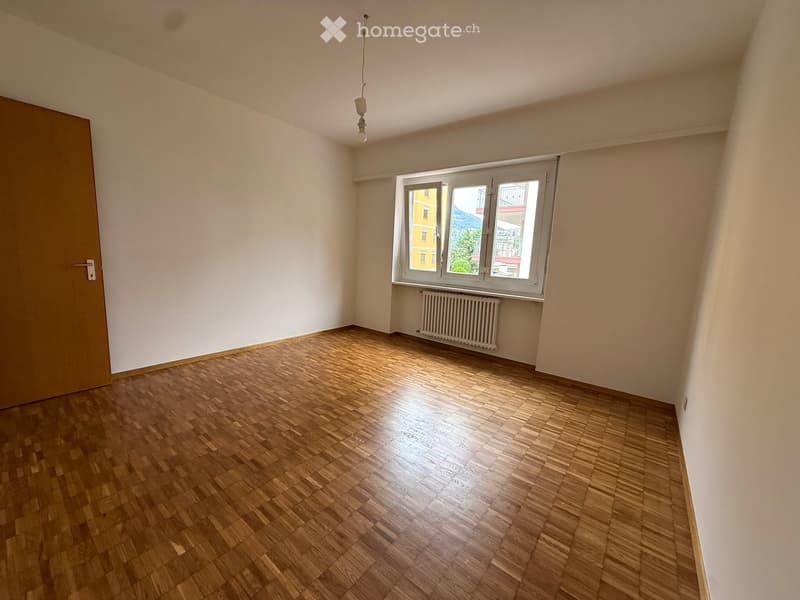 Empty room with wooden flooring, white walls, a window with white frame, a radiator, and a door with a silver handle.