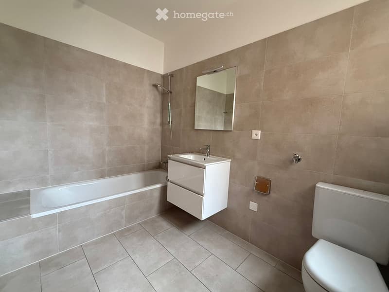Modern bathroom with a bathtub, sink, toilet, and a mirror above the sink. The walls are tiled in a neutral gray color, and the floor is tiled with matching tiles.