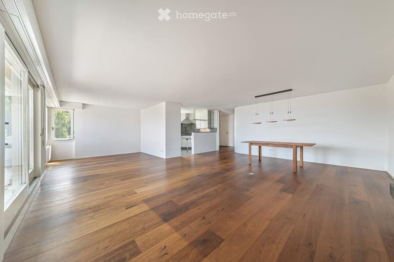 Bright, open living space with wooden flooring, a central wooden table, and a modern kitchen area in the background.