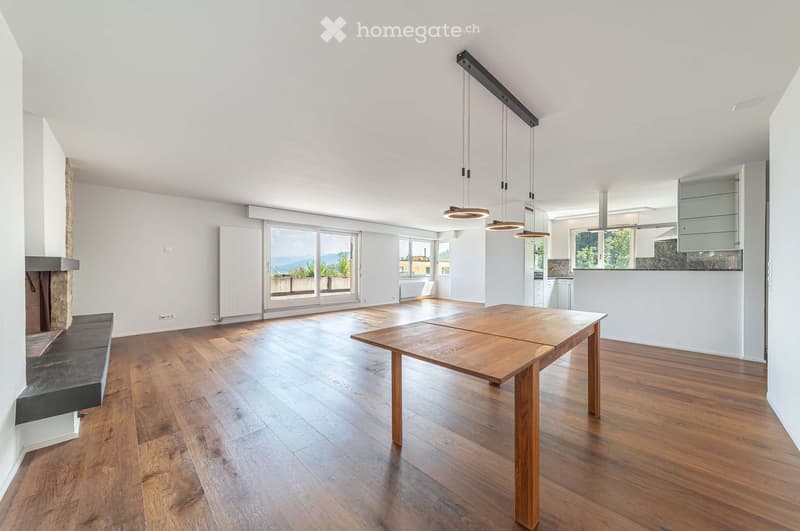Bright, open living space with wooden flooring, large windows offering a scenic view, a central wooden table, a stone fireplace with a bench, and modern pendant lighting.
