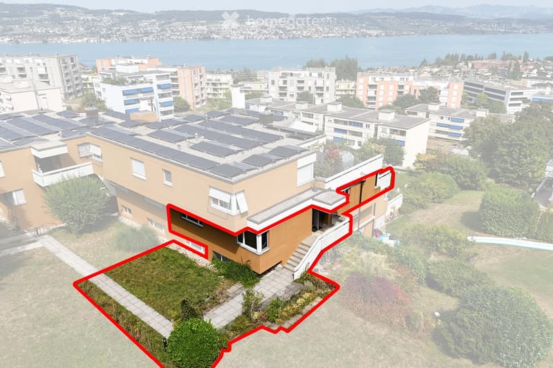 Aerial view of a multi-story residential building with solar panels on the roof, surrounded by greenery and neighboring structures, with a scenic lake and distant mountains in the background.