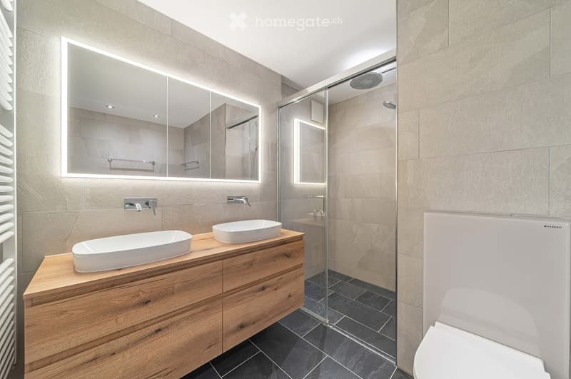 Modern bathroom with two sinks on a wooden vanity, a large mirror with LED lighting, a glass-enclosed shower, and a toilet.