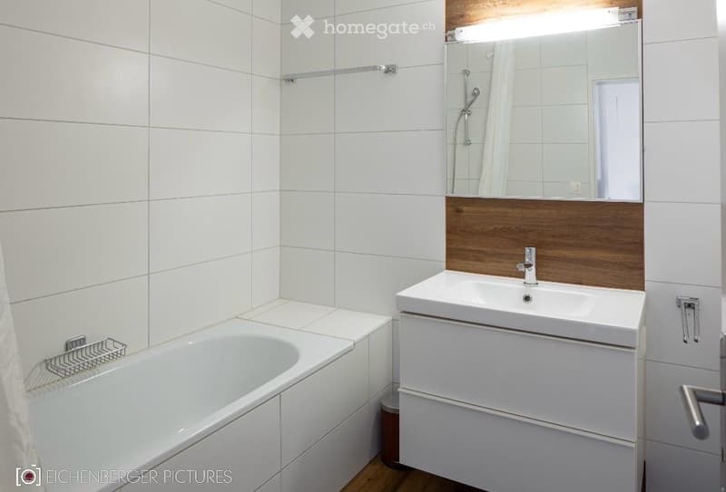 Modern bathroom with white tiled walls, a bathtub, a sink with a wooden backsplash, and a large mirror above the sink.