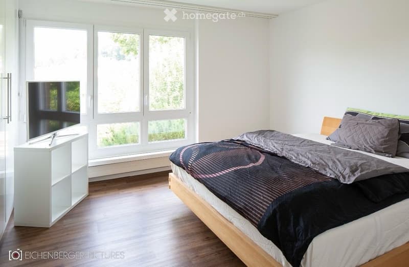 Modern bedroom with large windows offering a view, a wooden bed with a dark bedspread and pillows, a white nightstand, and a flat-screen TV on a white stand.