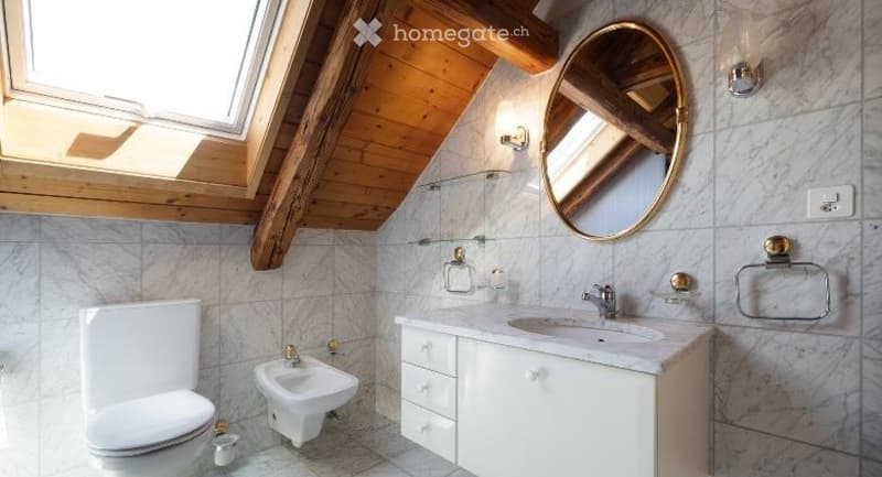 Modern bathroom with white tiles, wooden ceiling beams, and a skylight. Features a toilet, bidet, sink with cabinet, and a round gold-framed mirror.