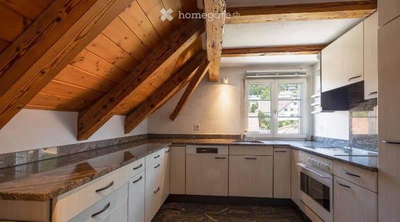 Bright kitchen with wooden beams, white cabinets, granite countertops, and a window overlooking the street.