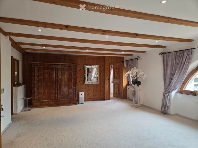 Spacious, well-lit room with wooden beams, a large wardrobe, a mirror, a small table with a vase, and curtains on the windows.